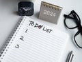 Handwriting text and list numbers on blank notepad with 2024 grey desk calendar standing with clock, pen, glasses.