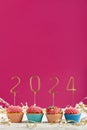 New Years background. Holiday cupcakes with numbers 2024 surrounded by New Years tinsel on pink background. Vertical frame. Copy