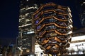 Vessel TKA, a spiral endless staircase, skyscrappers behind. Night view with bright lights. Hudson Yards, Manhattan`s West Side