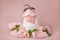 Newborn baby girl in a white cocoon on a pink background with pink roses and a hat like a flower