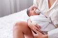 Newborn baby with teat look at mother who hold her and sit on the bed with day light