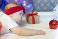 newborn girl in a red skirt and a red bow on her head lies on bed with christmas gift and ball against background of Royalty Free Stock Photo