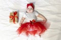 newborn girl in a red skirt and a red bow on her head lies on bed with christmas gift with red ribbon on a white Royalty Free Stock Photo