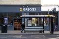 Exterior shot of Greggs Bakery showing company logo, signage and branding with a bus stop in front