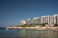 Newly built skyscrapers on the coast of Malta are reflected in the sea. The sky is blue. The sun is shining. People sunbathe on