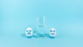 Next to the half-filled glass, two eggs with a cheerful and sad expression. Concept of optimist and pessimist. Blue background