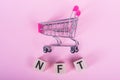 NFT - non-fungible token, word on wooden cubes, on a pink background with a shopping trolley