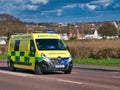 An NHS ambulance travelling at speed on a road in a semi-urban area of England, UK