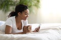 Nice App. Smiling Black Woman Using Smartphone And Drinking Coffee In Bed Royalty Free Stock Photo