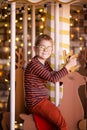 Nice blonde boy with glasses on the new year carousel with wooden deer and bright lights