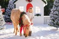 Nice blonde curly child and adorable pony with festive wreath near the small wooden house and snow-covered trees. New Year and Chr Royalty Free Stock Photo