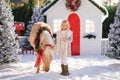 Nice blonde curly child and adorable pony with festive wreath near the small wooden house and snow-covered trees. New Year and Chr Royalty Free Stock Photo