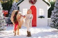 Nice blonde curly child and adorable pony with festive wreath near the small wooden house and snow-covered trees. New Year and Chr Royalty Free Stock Photo