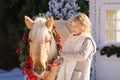 Nice blonde curly child and adorable pony with festive wreath near the small wooden house and snow-covered trees. Royalty Free Stock Photo