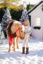 Nice blonde curly child caresses adorable pony with festive wreath near the small wooden house and snow-covered trees. New Year an Royalty Free Stock Photo