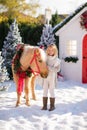 Nice blonde curly child caresses adorable pony with festive wreath near the small wooden house and snow-covered trees. New Year an Royalty Free Stock Photo