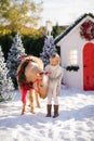 Nice blonde curly child caresses adorable pony with festive wreath near the small wooden house and snow-covered trees. New Year an Royalty Free Stock Photo