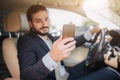 Nice and cheerful businessman sits in luxury car. He holds phone in one hand and looks at its screen. Another hand is on Royalty Free Stock Photo