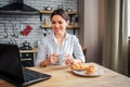 Nice cheerful businesswoman sit at table in kitchen and look at laptop. She works at home. Model hold white cup with