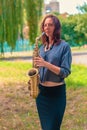 Nice girl with red hair in a blue sweater and black skirt is standing with a yellow saxophone in the forest Royalty Free Stock Photo