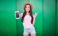 Nice girl stands and poses on camera. She holds white phone in hand and showes piece symbol wih the other one. Woman is