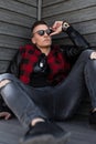 Nice handsome young hipster man in black sunglasses in a trendy red checkered jacket in sneakers in gray ripped jeans is sitting