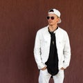 Nice hipster young man in a trendy white summer jacket in a stylish black t-shirt in a fashionable baseball cap in dark sunglasses