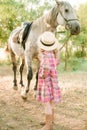 A nice little girl with light curly hair in a vintage plaid dress and a straw hat and a gray horse. Horses a
