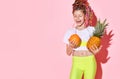 Nice little girl with multicolored cornrows on head laughing joyfully with closed eyes holding pineapple and orange in hands Royalty Free Stock Photo