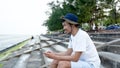 A nice-looking Asian guy wearing his favorite hat, sitting on a chair and enjoying the sea view on the beach, Royalty Free Stock Photo