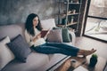 Nice-looking attractive lovely focused calm peaceful wavy-haired girl sitting on divan reading book long crossed legs