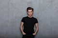 Nice modern young man hipster in a black fashionable t-shirt in jeans posing near a vintage wall indoors. Handsome guy model in Royalty Free Stock Photo