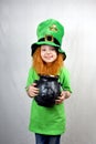 Nice smiling small girl with decorative red beard, green clover leaf on her cheek and leprechaun hat, holds cast iron pot, full of Royalty Free Stock Photo