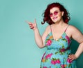 Nice smiling to us plus-size overweight lady in sunglasses and sundress point her finger at something on text copy space on mint