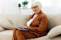 a nice, sweet elderly woman is sitting on the sofa in a bright apartment typing text in a laptop holding it on her lap