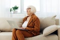 a nice, sweet elderly woman is sitting on the sofa in a bright apartment typing text in a laptop holding it on her lap