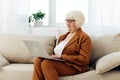 a nice, sweet elderly woman is sitting on the sofa in a bright apartment typing text in a laptop holding it on her lap