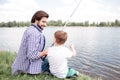Nice view of happy son and dad sitting together at river shore. Guy is looking at his son and fishing. Boy is looking at Royalty Free Stock Photo