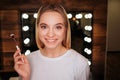 Nice young blonde woman look on camera and smile. She stand in beauty room. Woman hold brush for make up. Mirror with Royalty Free Stock Photo