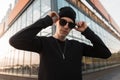 Nice young hipster man in a black shirt in a knitted fashionable hat straightens dark trendy sunglasses and looks into the camera Royalty Free Stock Photo