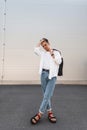 Nice young man in a stylish white shirt in blue fashionable jeans with a black vintage bag poses in the street on a sunny day. Royalty Free Stock Photo