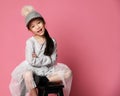 Nicely smiling asian fashion kid girl in pink dress and funny cap with fur pompon