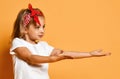 Nice kid girl in white t-shirt and red headband is holding something on her open palms on yellow with copy space