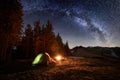 Night camping. Illuminated tent and campfire near forest under night sky full of stars and milky way
