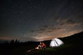 Tourists near campfire and tent under night starry sky