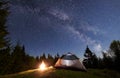 Night camping in mountains. Tourist tent by campfire near forest under blue starry sky, Milky way