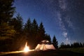 Night camping in mountains. Tourist tent by campfire near forest under blue starry sky, Milky way Royalty Free Stock Photo