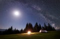 Night camping in mountains. Tourist tent by campfire near forest under blue starry sky, Milky way Royalty Free Stock Photo