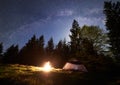 Night camping in mountains. Tourist tent by campfire near forest under blue starry sky, Milky way Royalty Free Stock Photo