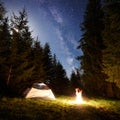 Night camping in mountains. Tourist tent by campfire near forest under blue starry sky, Milky way Royalty Free Stock Photo